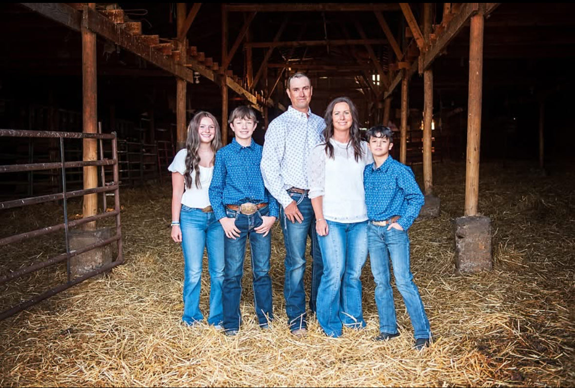 Family of five posing in a barn with wooden beams and hay on the floor.