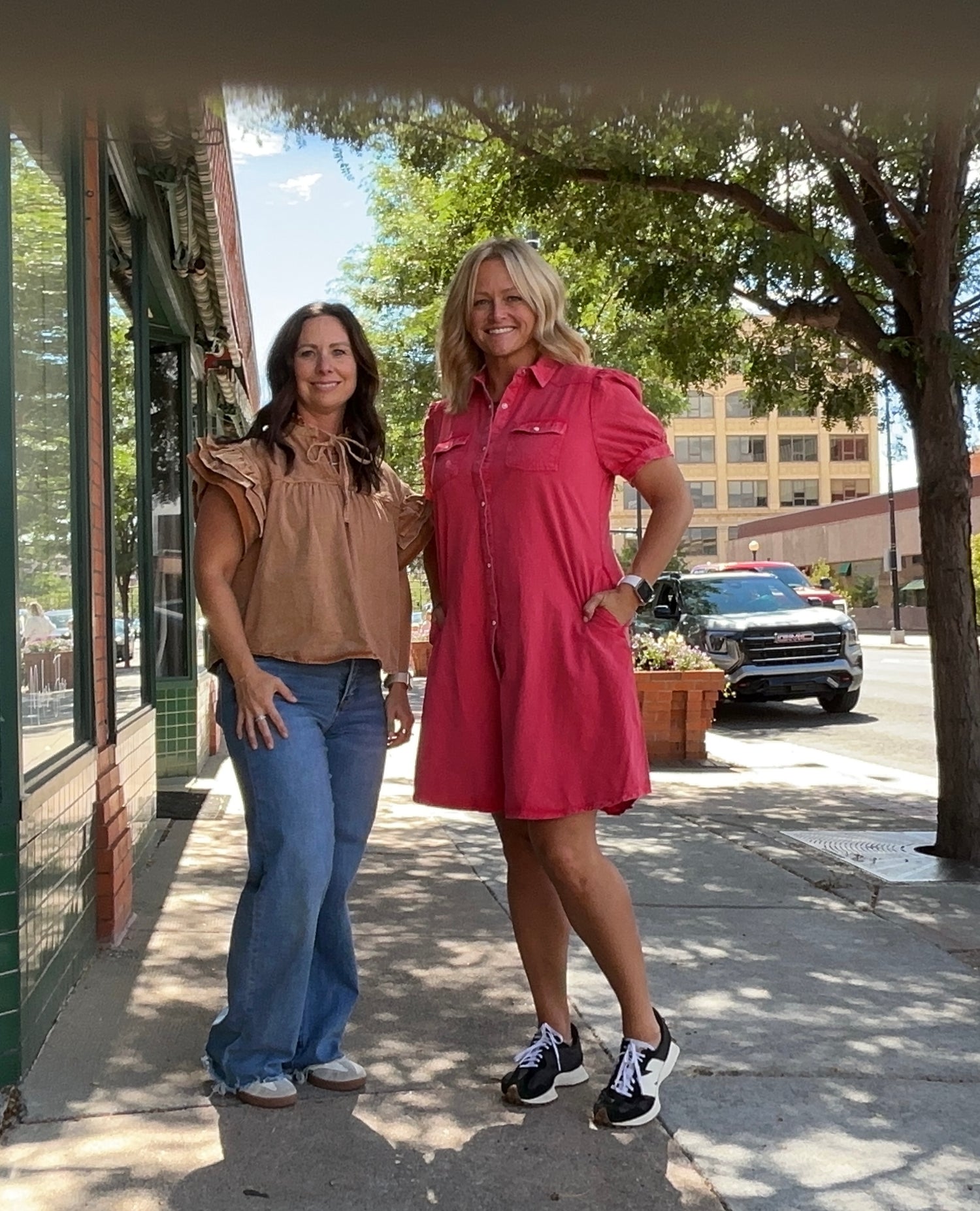 Two women standing on a sidewalk with a building and tree in the background