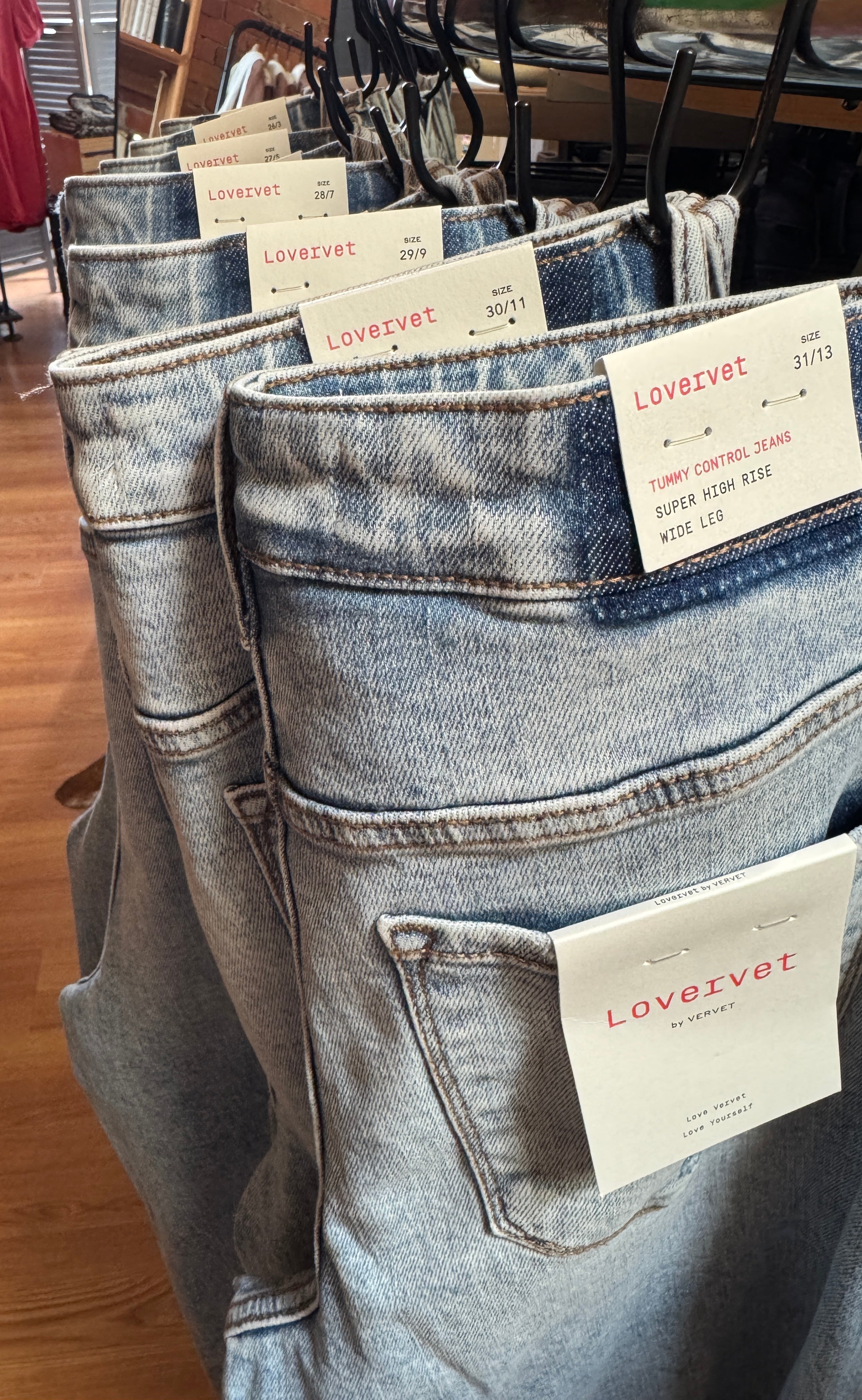 Stack of blue jeans with tags on a wooden floor, likely in a store setting.