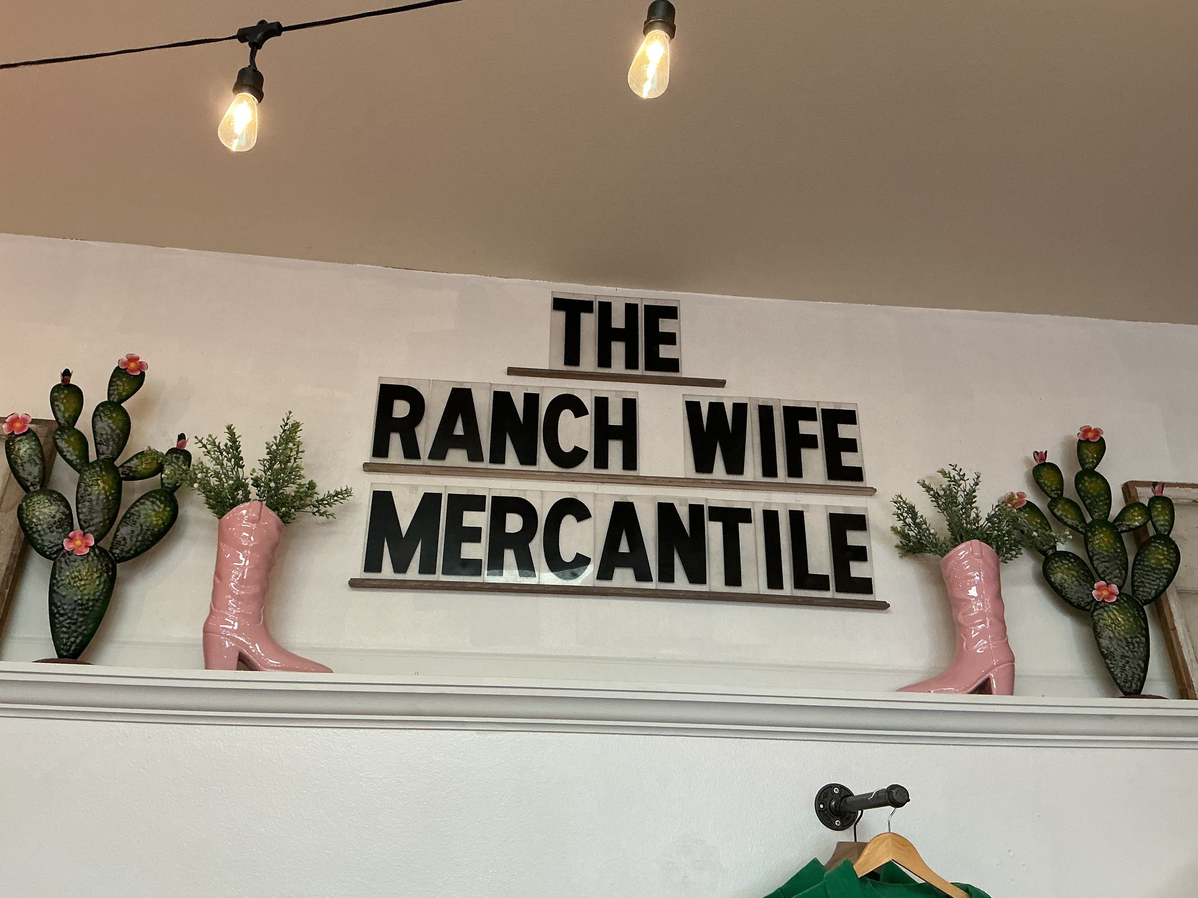 Decorative sign with 'The Ranch Wife Mercantile' on a wall with cacti and boots.